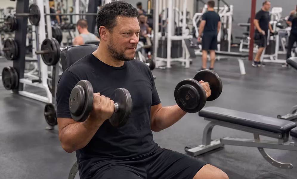 A man seated on a gym bench curling a pair of dumbbells.